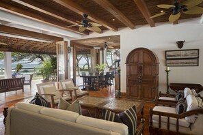A spacious living area with a wooden ceiling, large windows, and a view of the sea. There are multiple seating arrangements including a sofa, chairs, and a dining table. - Casa Encantos Beach Front House (Hacienda Iguana)