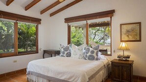 Ein Schlafzimmer mit einem Bett, zwei Fenstern, einer Holzbalkendecke, einem Nachttisch und einer Lampe.