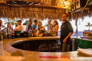 A bar area with a thatched roof, a counter with various bottles and glasses, and a view of palm trees and the ocean.