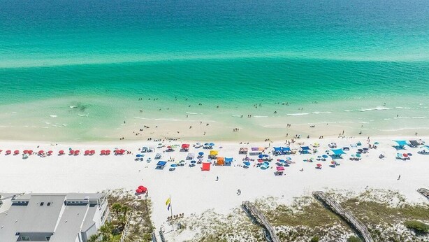 Une vue aérienne d’une plage, avec de nombreux parasols colorés et des chaises de plage, une mer bleue et limpide, et une rive sablonneuse.