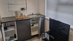 A compact kitchen with a built-in sink, a glass shelf with cups and glasses, and a black TV stand.