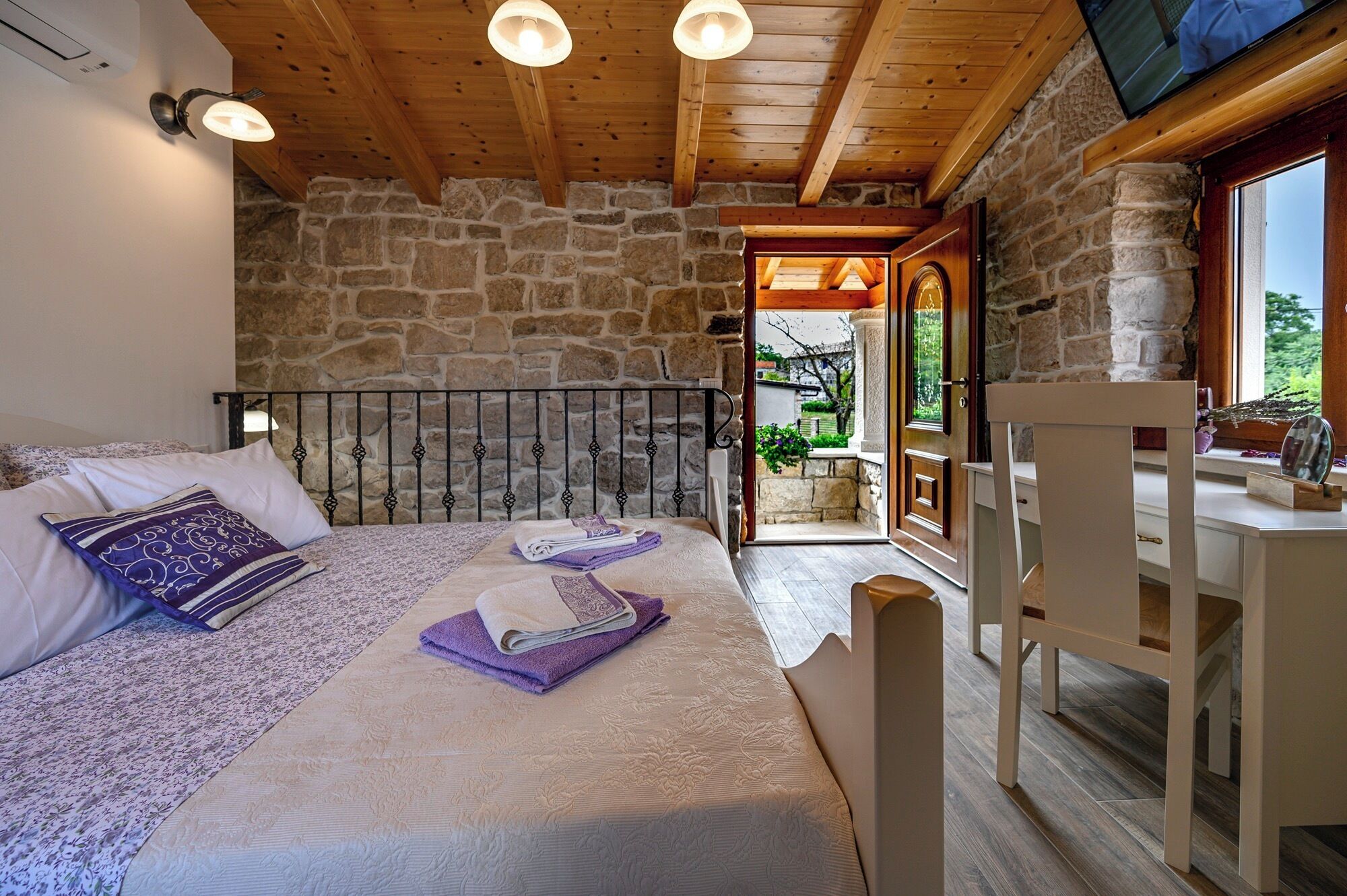 Ein Schlafzimmer mit Steingemäuer, einem Bett mit Kopfteil, Holzboden und Blick ins Freie durch eine Tür.