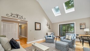 A spacious living room with a high ceiling, a sofa, two armchairs, a coffee table, and a dining area with a table and chairs. There are framed pictures on the walls and a window with a view of trees. - LABOR DAY SPECIAL 2 NIGHTS FREE Lake Winnipesaukee Waterfront Sleeps 14 (Moultonborough)
