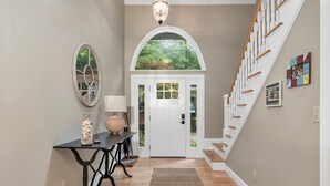 A hallway with a wooden staircase, a white door, a round mirror, a table with a lamp, and a rug. - LABOR DAY SPECIAL 2 NIGHTS FREE Lake Winnipesaukee Waterfront Sleeps 14 (Moultonborough)