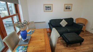 Un salon avec un canapé gris foncé, une table basse en bois et un vase bleu contenant des fleurs séchées. Deux tableaux encadrés ornent le mur, et une fenêtre offre une vue sur une voiture garée à l’extérieur.
