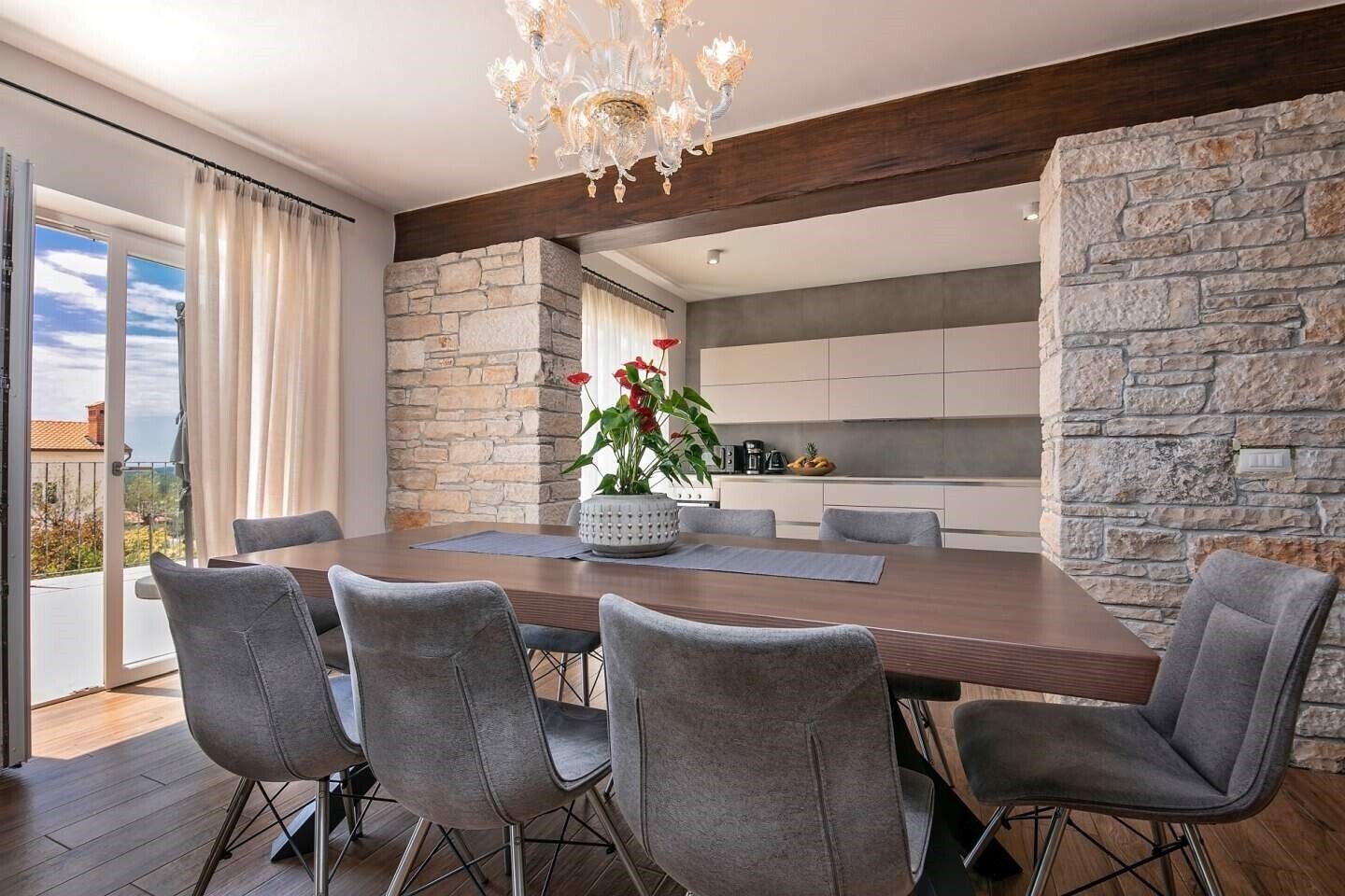 A modern dining area with a large wooden table, grey upholstered chairs, a decorative chandelier, and a view of the outdoors through large windows.