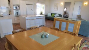 A modern kitchen with white appliances, a dining area with a wooden table and chairs, and a living room with a sofa and a coffee table.