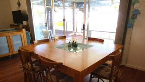 A dining area with a wooden table and chairs, a glass-top table, and a view through large windows.