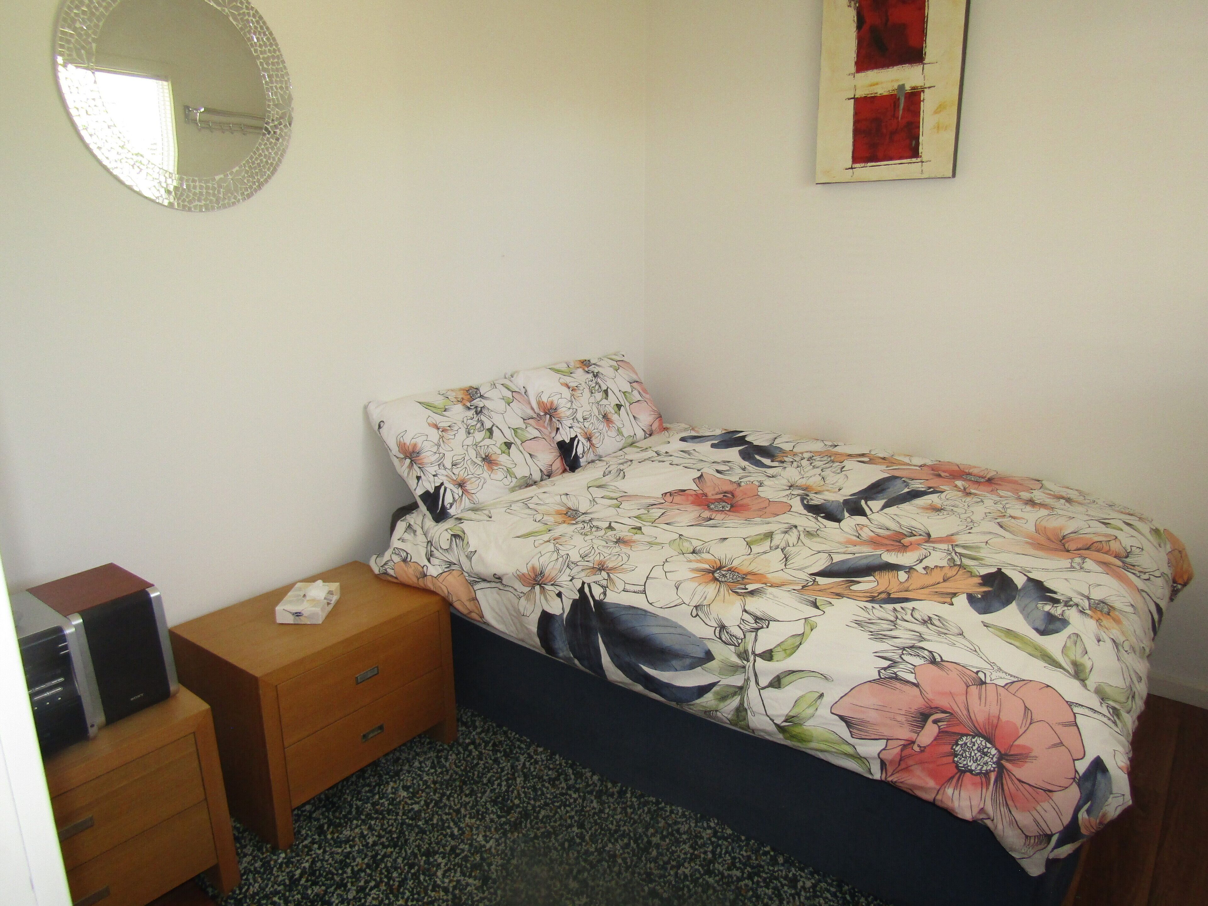 A bedroom with a floral bedspread, a bedside table with a lamp, a small wooden nightstand, and a mirror on the wall.