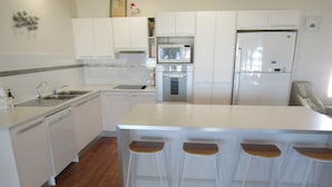A modern kitchen with white cabinetry, a stainless steel refrigerator, a microwave, and a dishwasher. There are four bar stools with wooden seats.