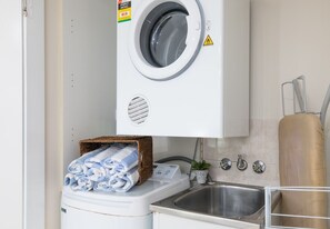 A compact laundry area with a front-loading washing machine, a small sink, and a basket of folded towels. - Airlie Horizon (Airlie Beach)