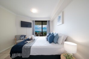A hotel room with a bed, a TV mounted on the wall, a window with a view, and a bedside table with a lamp. - Airlie Horizon (Airlie Beach)