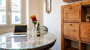 A round glass table with a laptop, a bottle of wine, and a glass. There is a wooden cabinet with drawers labeled "ARGENTINA" and "LUP." A mirror hangs on the wall, and a window is visible in the background. - Borgo Ognissanti Suites Stay in the Heart of Florence! 🔝 brand new location (Firenze)