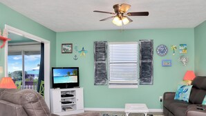 Un salon avec un ventilateur de plafond, un téléviseur, un canapé, une table basse et une fenêtre avec des volets.
