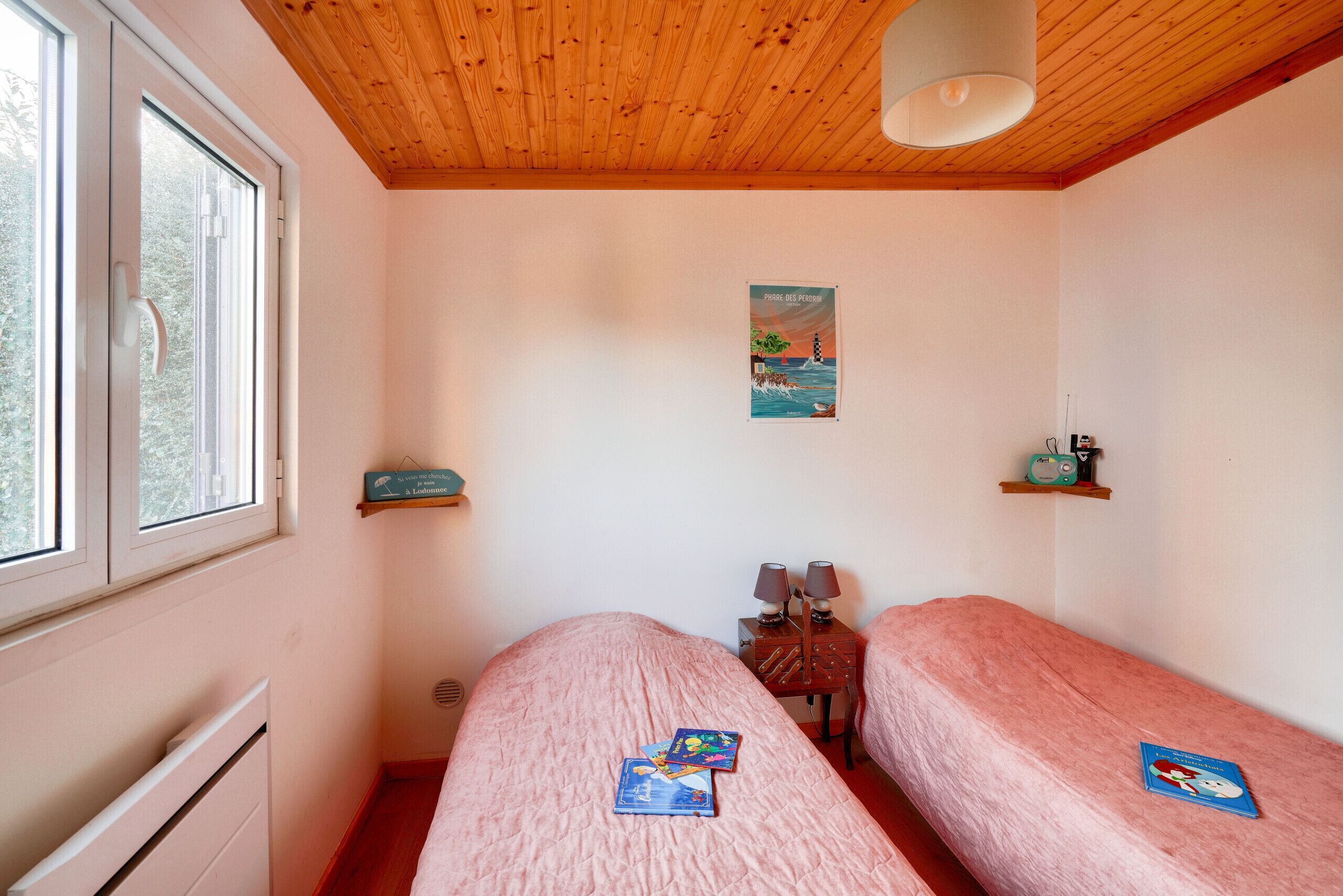 Een slaapkamer met twee enkele bedden, een houten plafond, een raam, een radiator en een klein tafeltje met lampen.