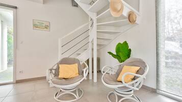 Een modern interieur met een witte spiraaltrap, twee rieten fauteuils met kussens, een grote groene plant en een ingelijst kunstwerk aan de muur.