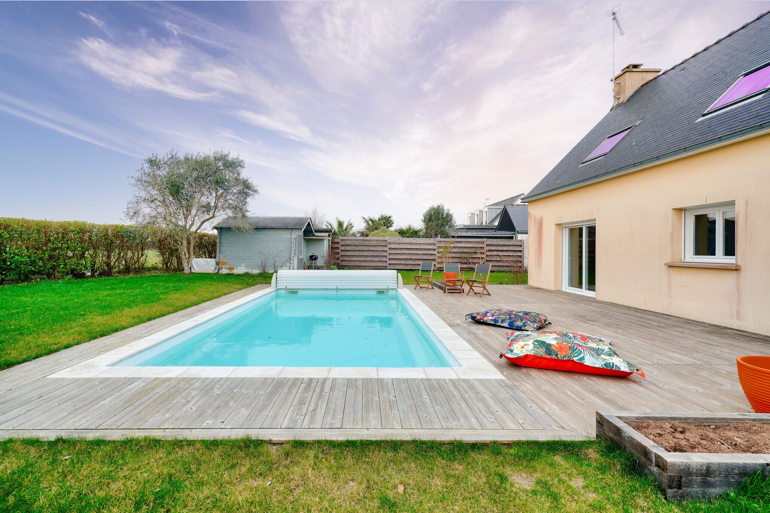 En rektangulær swimmingpool med en træterrasse, græsplæne og et hus med skråt tag og vinduer.