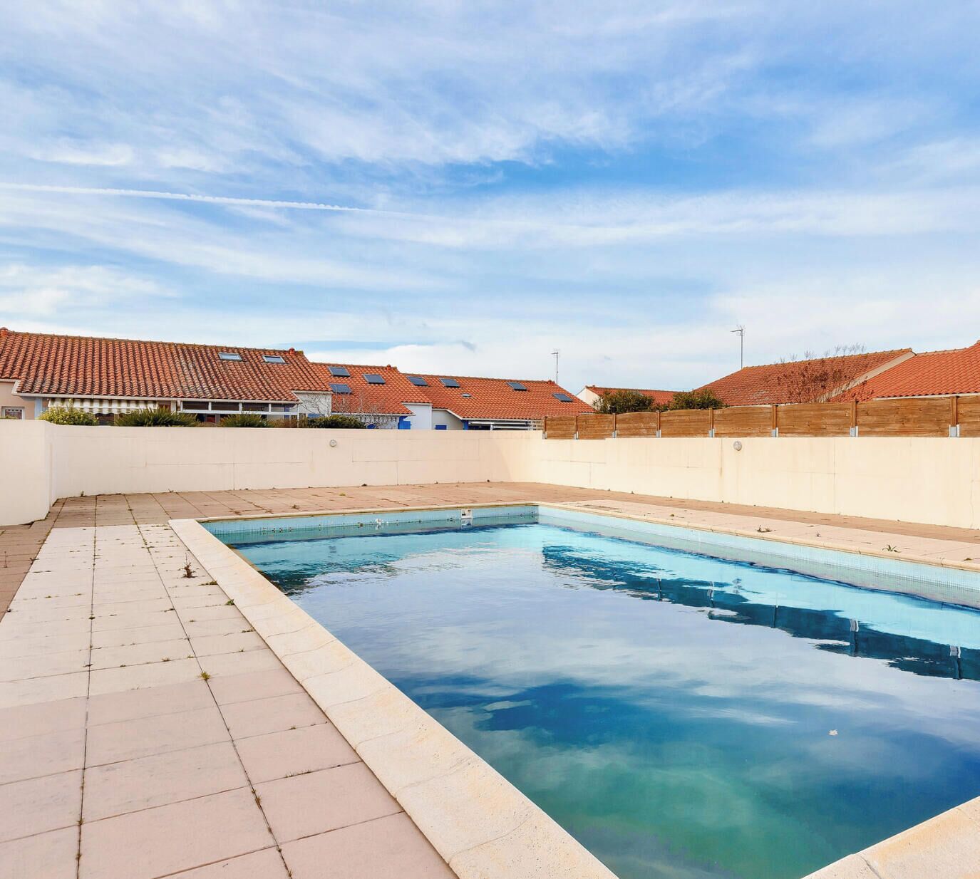 Une piscine dotée d’une terrasse carrelée, entourée d’un petit muret et bordée d’un bâtiment dont le toit est également recouvert de tuiles.