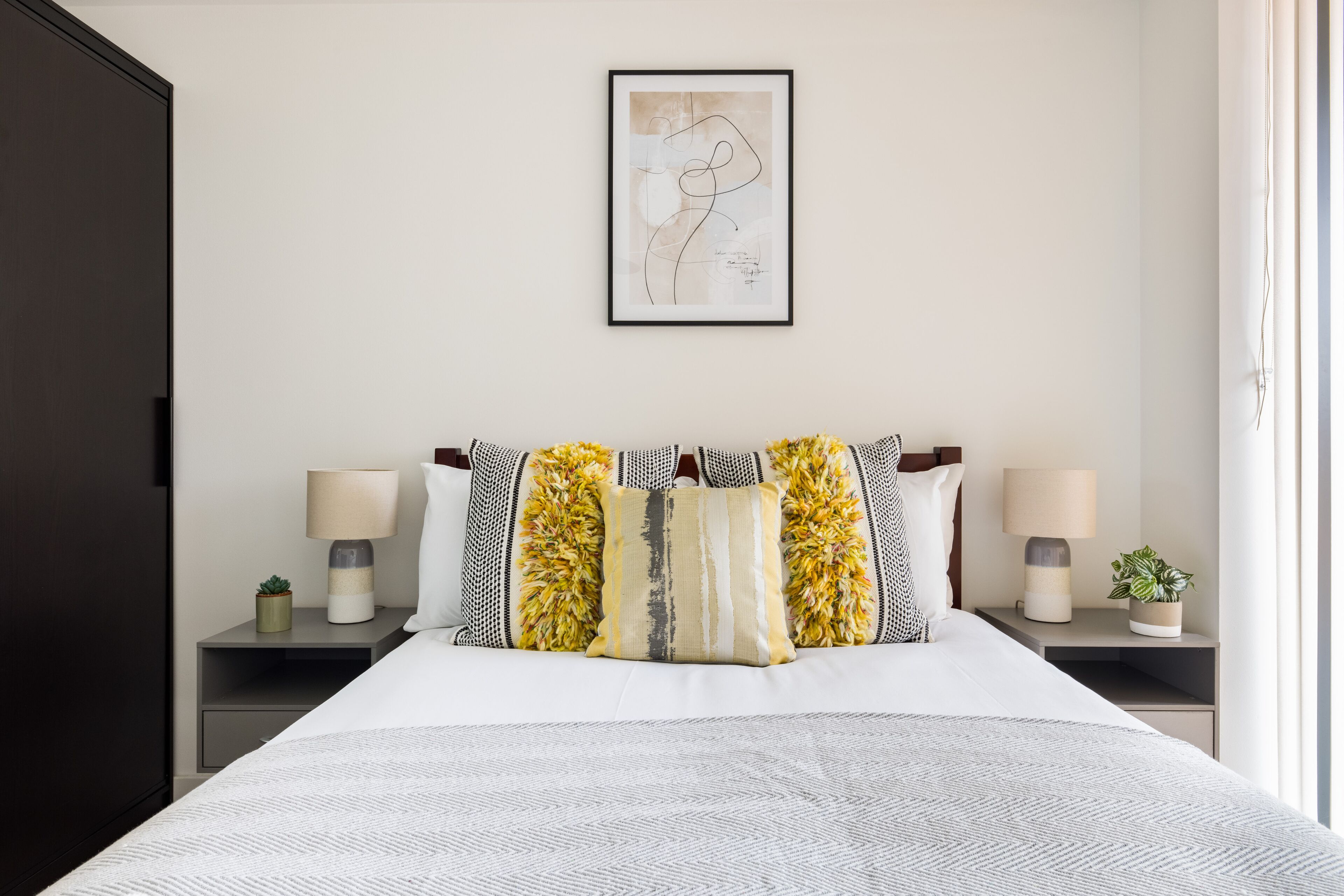 A neatly arranged bedroom with a large bed, two bedside tables with lamps, a framed artwork on the wall, and a potted plant on one of the nightstands.