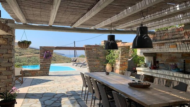 An outdoor patio with a stone floor, a long table, wicker chairs, hanging black lamps, and a stone wall with a hanging planter.
