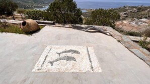 A tiled patio with a mosaic of a dolphin, a large cylindrical object, and a view of the sea and distant buildings. - Holiday House - Koundouros Panoramic Sea View (Kea)