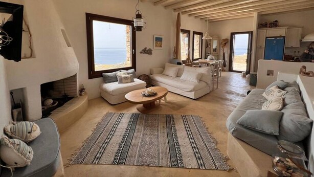 A spacious living room with a large window overlooking the sea, a fireplace, and a kitchen area visible through an open door. - Holiday House - Koundouros Panoramic Sea View (Kea)