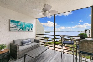 A balcony with a view of a body of water and a bridge, furnished with a sofa, a table, and chairs.
