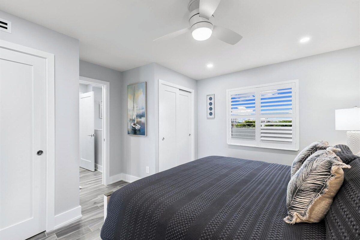 Une chambre à coucher avec un lit, un ventilateur de plafond, une fenêtre avec des stores et une porte.