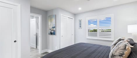 Quarto com cama, ventoinha no teto, janela com persianas e porta.