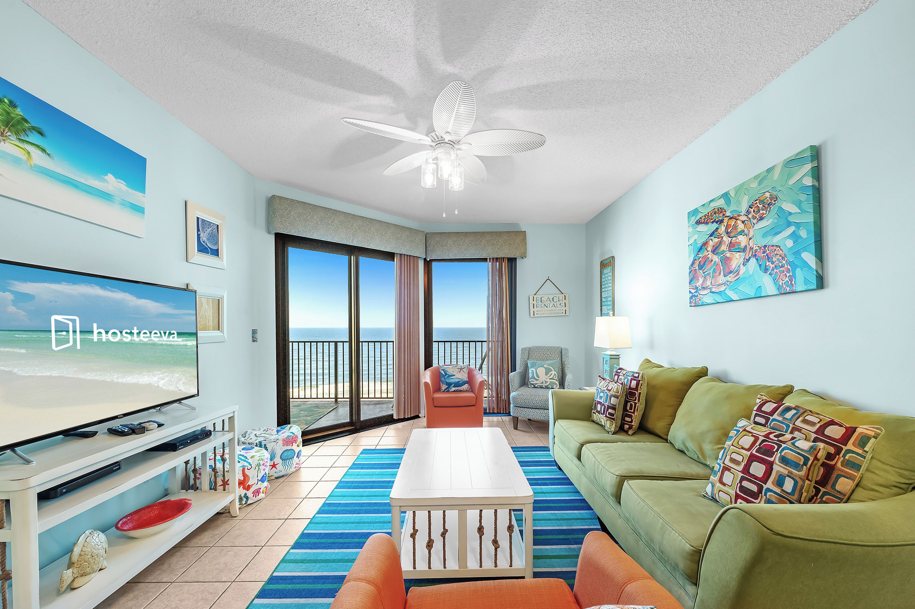 A living room with a TV, a ceiling fan, a sofa, a coffee table, and a view of the ocean through large windows.