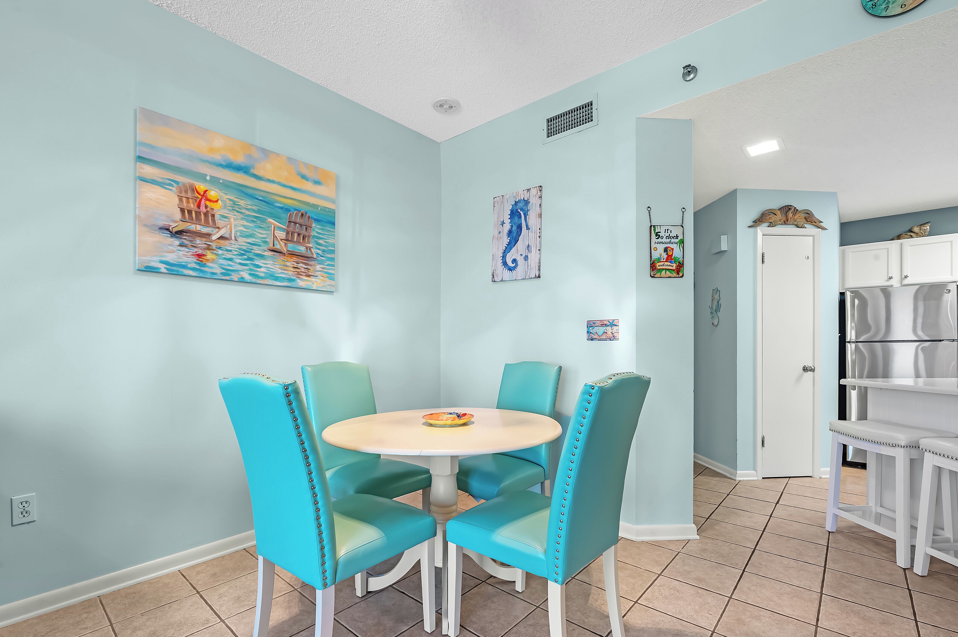 A dining area with turquoise chairs and a white table, a painting of a boat on water, and a door leading to a kitchen with white cabinets.