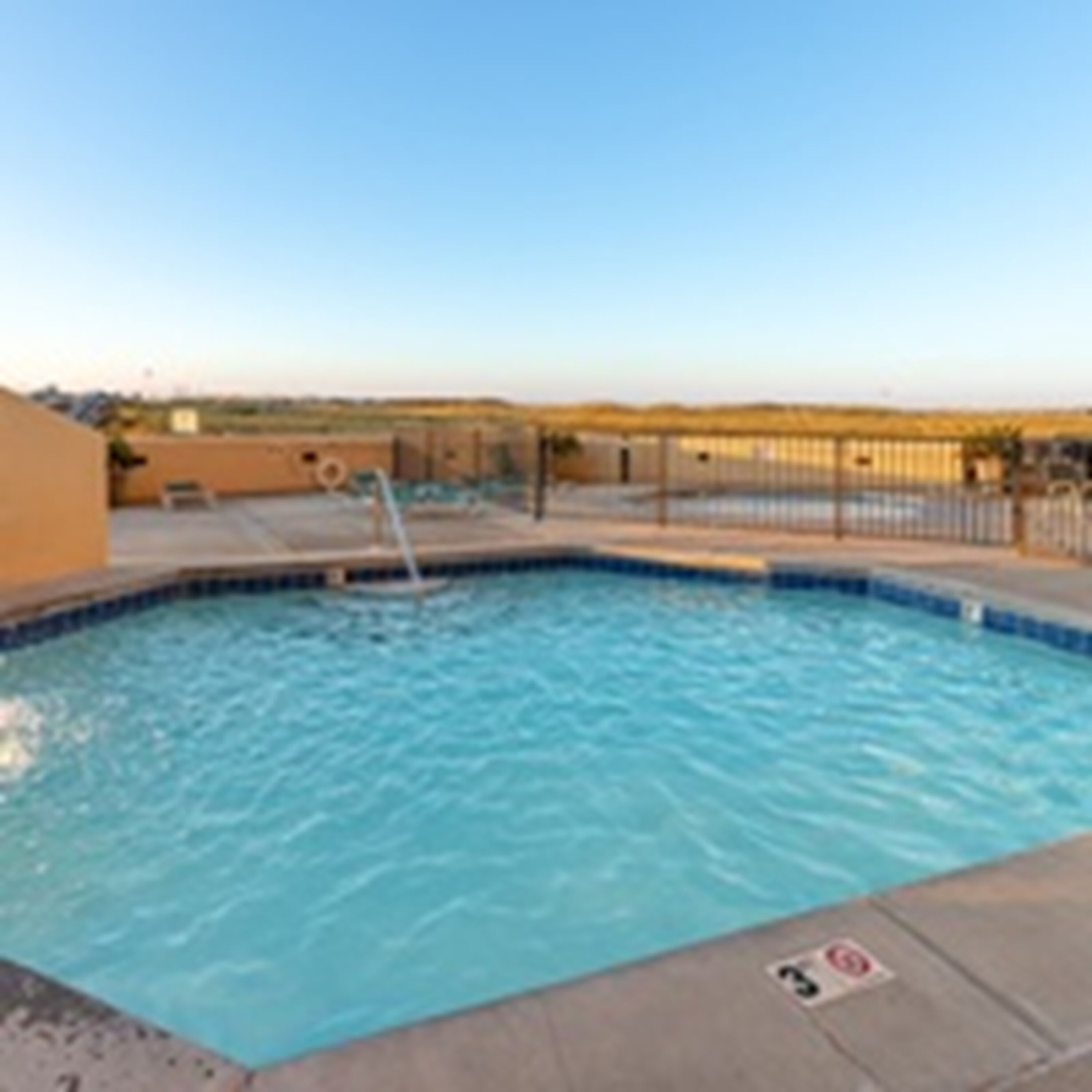 A swimming pool with a hot tub, surrounded by a fence and a clear sky.
