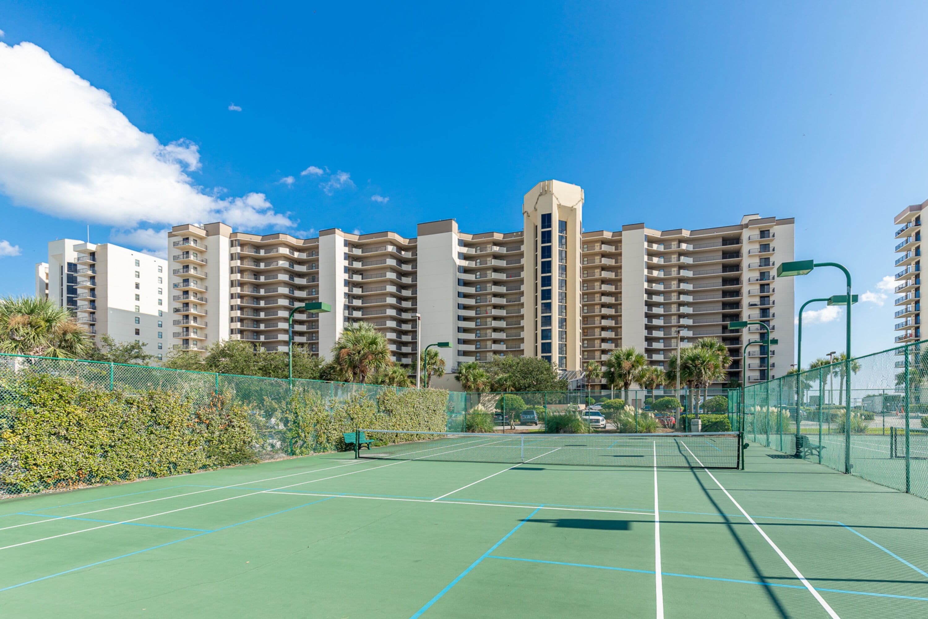 A tennis court with a net, surrounded by a fence and greenery.