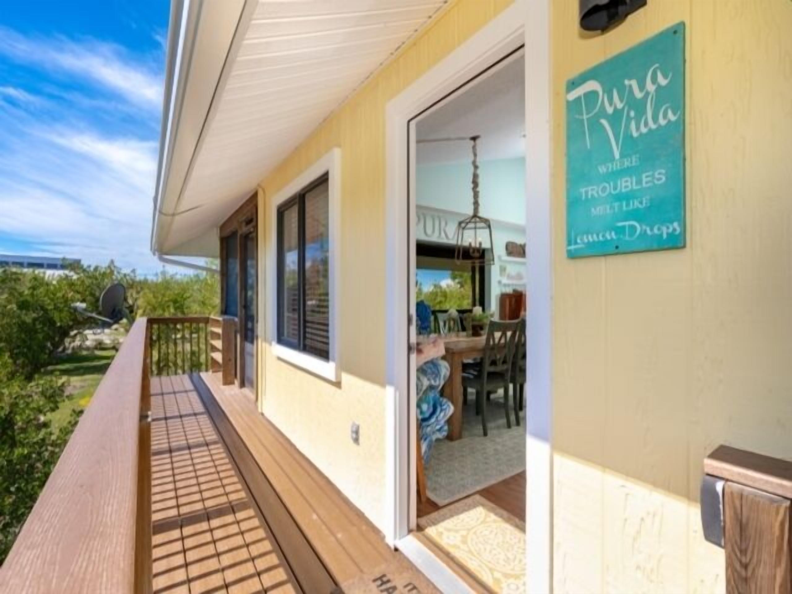 Eine Holzterrasse mit einer Glastür, die zu einem Essbereich führt. Ein Schild an der Wand zeigt: 'Pura Vida Where Troubles Melt Like Lemon Drops'.