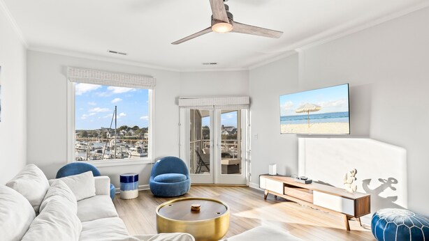 A modern living room with a large window offering a view of the beach and marina, a ceiling fan, and a flat-screen TV mounted on the wall. - Moorings 510 - Yacht View Serenity! Top Floor! (Isle of Palms)