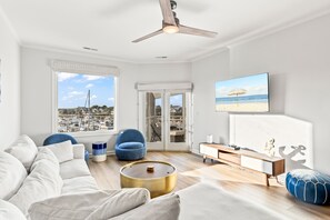 A modern living room with a large window offering a view of the beach and marina, a ceiling fan, and a flat-screen TV mounted on the wall. - Moorings 510 - Yacht View Serenity! Top Floor! (Isle of Palms)