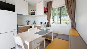 Cozinha compacta com armários brancos, micro-ondas e cafeteira. Há uma mesa de jantar com cadeiras e um banco. Uma janela com cortinas é visível.