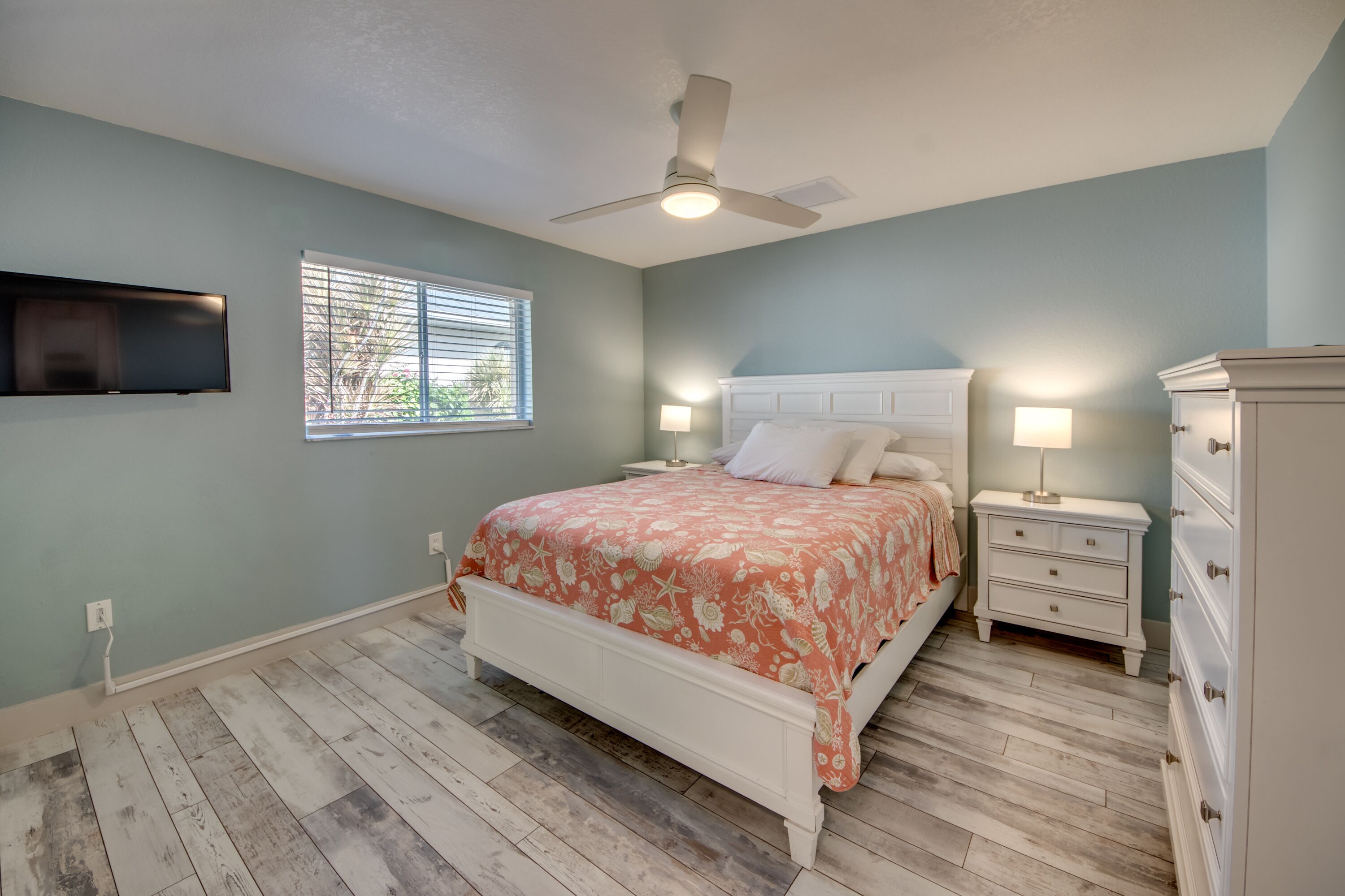 A bedroom with a bed, nightstands, a dresser, a ceiling fan, and a TV mounted on the wall.