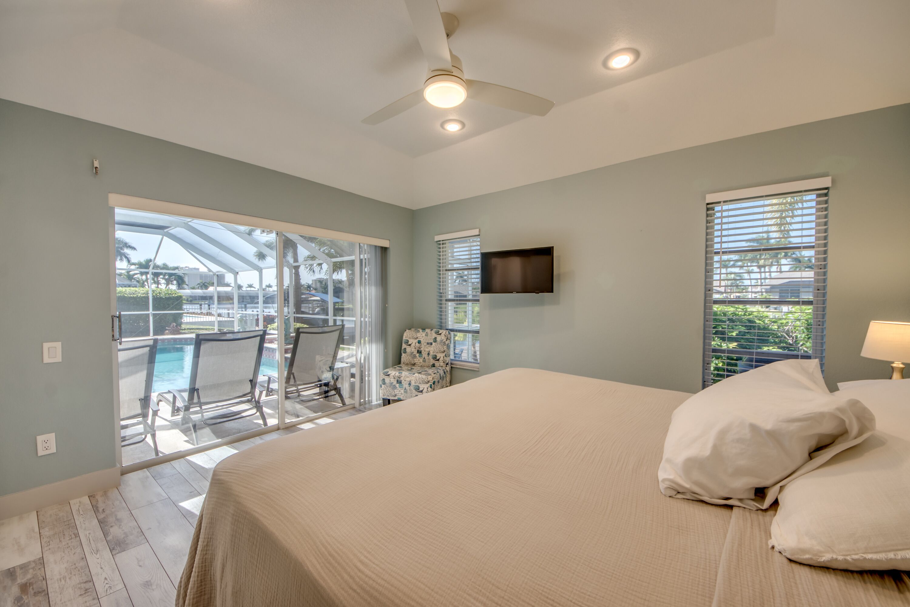 Ein Schlafzimmer mit einem großen Bett, einem Deckenventilator, einem an der Wand montierten Fernseher und Blick auf einen Pool und eine Außenlounge durch eine Schiebetür.