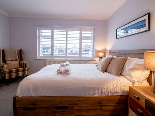 A neatly made bed with rolled towels, a wooden headboard, a chair with a cushion, a bedside lamp, and a window with blinds.