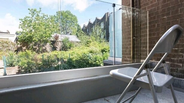 A balcony with a folding chair, a glass door, and a view of greenery and buildings. - Deluxe Central London North Apartment (Greater London)