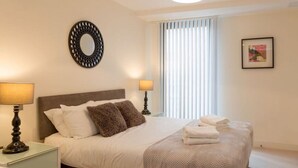 A neatly arranged bedroom with a bed, two bedside lamps, a round mirror, a window with blinds, and a framed picture on the wall. - Capacity House · Central Tower Bridge Apartment (London)