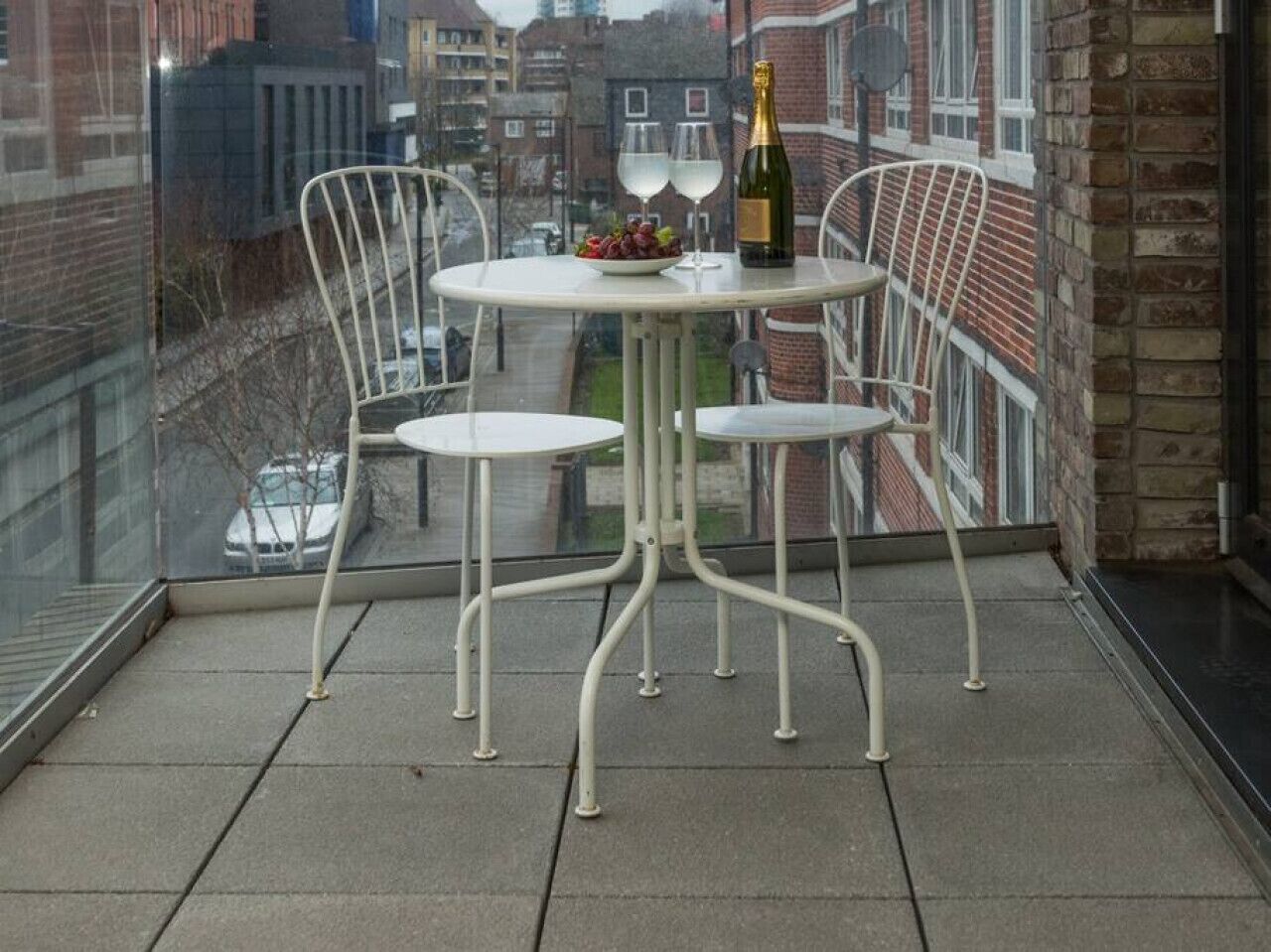 En balkon med et rundt bord, to hvide stole, en flaske champagne og en skål med frugt.