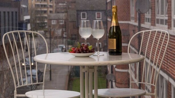 A round table with two white chairs, a bottle of champagne, two glasses, and a bowl of grapes. - Capacity House · Central Tower Bridge Apartment (London)