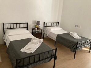 A room with two single beds, each with a dark green quilt and white linens. There is a small wooden nightstand with a lamp and a potted plant between the beds.