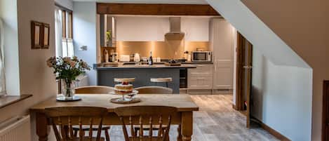 Un área de comedor con una mesa y sillas de madera, un jarrón con flores, y una cocina al fondo con microondas y tostadora.