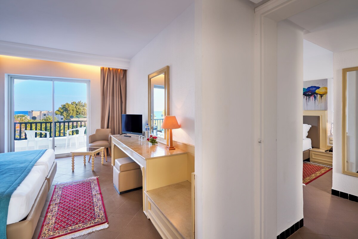 A hotel room with a bed, a desk, a TV, a balcony with a view of the sea, and a bathroom.