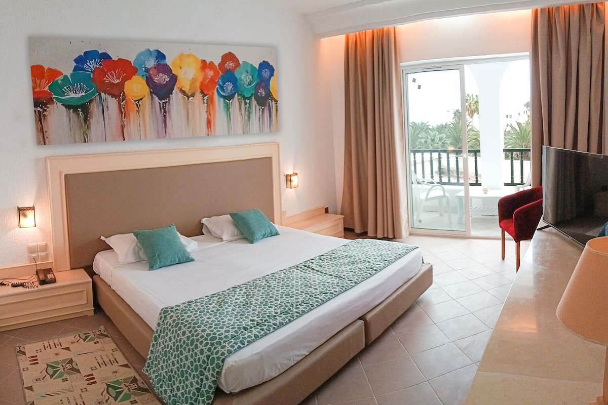 A hotel room with a large bed, a TV, a balcony with a chair, and a painting above the bed.