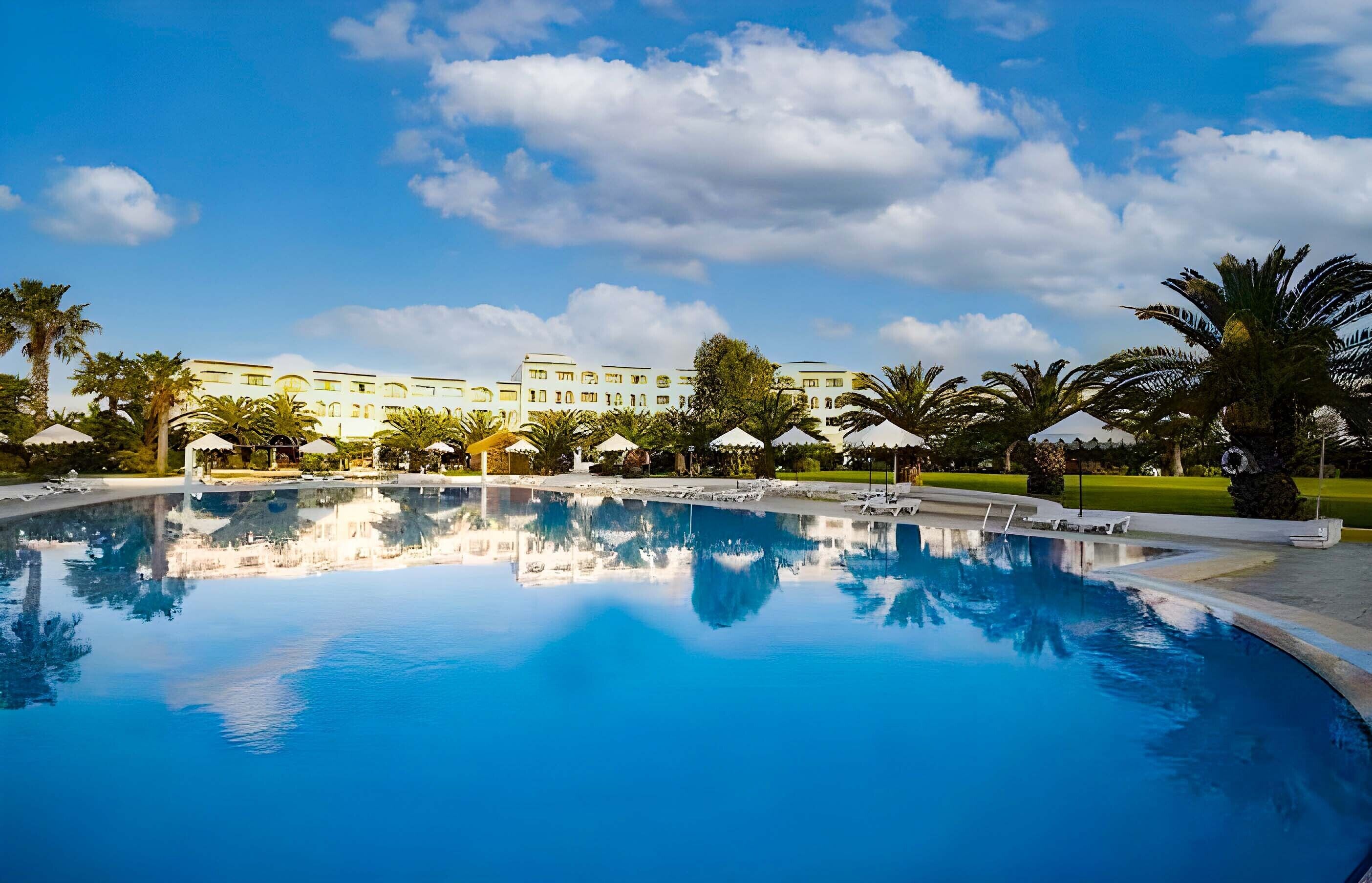 Una piscina que refleja palmeras y un hotel al fondo.