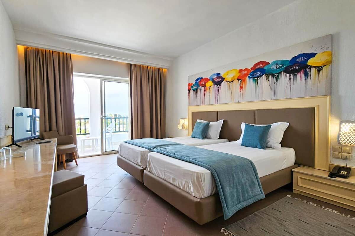 A hotel room with a large bed, a desk, a chair, a TV, and a balcony with curtains.